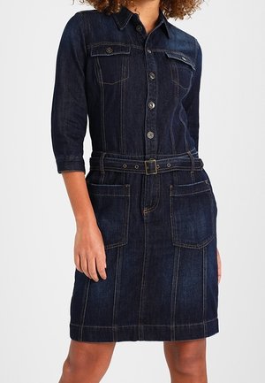 Kvinde iført en mørkeblå denimkjole med bælte, trekvartærmer og store forlommer.