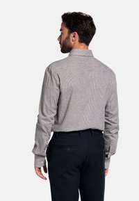 Man die met zijn rug naar de camera staat en een langwerpig geruit shirt draagt dat in een donkere op maat gemaakte broek is gestoken, tegen een witte achtergrond.