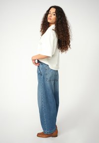 Loose-fitting white t-shirt, blue wide-leg jeans, and brown woven ankle boots. Curly hair falls past shoulders. Seam detailing on jeans.
