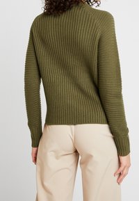Olivgrüner, gerippter Strickpullover mit langen Ärmeln und abgerundetem Saum, der aus strukturiertem Material und taillierten Bündchen besteht, kombiniert mit hellbeigen Hosen.