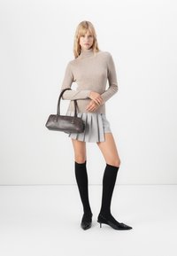 Kvinde står i en beige højhalset sweater, lys grå plisseret nederdel, sorte knælange strømper, sorte hæle og holder en brun håndtaske på den ene arm.