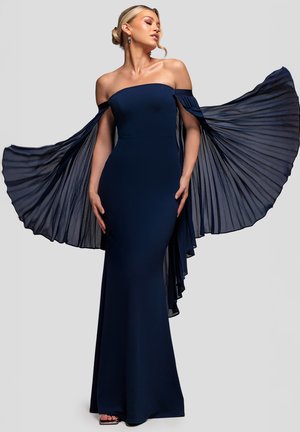 Donna in abito blu navy con scollo a barca e maniche mantella plissettate in tessuto trasparente, con le braccia distese, che guarda di lato su uno sfondo grigio chiaro.
