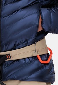Marineblauwe pufferjack met diagonale gestikte textuur, gecombineerd met een beige riem met een feloranje karabijnhaak. Ritsdetail zichtbaar.