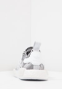 Svart och vitrandig atletisk sko med stickad ovandel, vit gummisula och bakre dragflik med logotyp. Texturerad design förbättrar greppet.