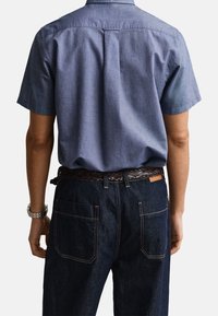 Homme portant une chemise bleue à manches courtes boutonnée rentrée dans un jean en denim foncé avec une ceinture tressée marron, vu de dos.