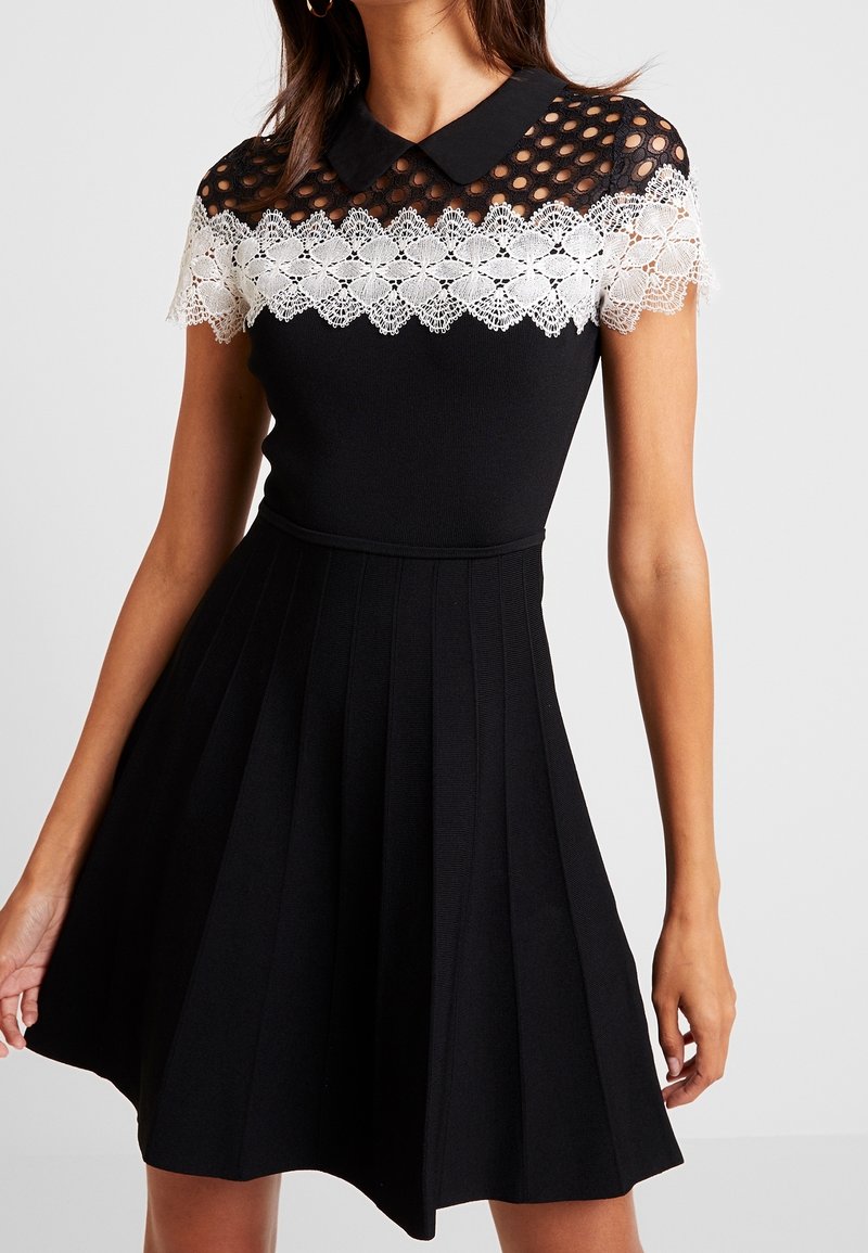 Schwarzes Kleid mit taillierter Oberpartie, plissétem Rock und Kragen. Spitzenakzente an den Schultern mit kreisförmigen Ausschnitten und floralen Mustern.