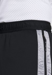 Svarta träningbyxor med midjeband i svart elastiskt material, med en sidoficka med dragkedja och en grå panel samt en texturerad, glänsande yta.