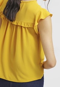 Blusa gialla con maniche a palloncino arricciate, tessuto testurizzato e vestibilità ampia, con un design posteriore arricciato.