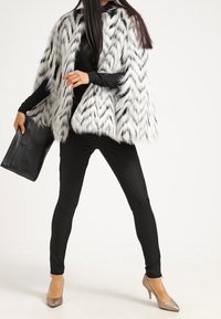 Capa de piel sintética con un patrón de zigzag en blanco y negro, combinada con pantalones negros ajustados y tacones metálicos plateados; sosteniendo un bolso de mano negro.