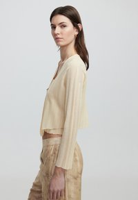Cardigan beige corto con costine verticali, maniche lunghe e scollatura a V. Indossato con pantaloni marrone chiaro testurizzati con motivi floreali.