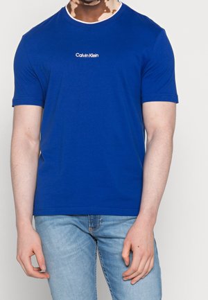 Mand iført en ensfarvet blå Calvin Klein t-shirt og lyseblå jeans står foran en hvid baggrund.