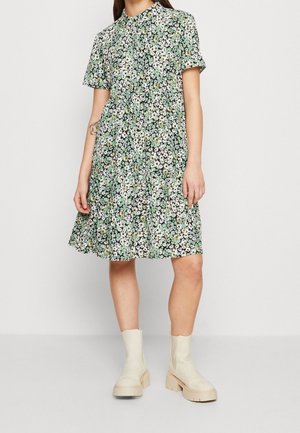 Vrouw draagt een kort mouw, knielange bloemenjurk in wit, groen en zwart, gecombineerd met crèmekleurige, chunky enkellaarzen.