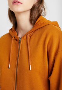Sudadera con capucha naranja y cremallera, con un tejido texturizado, cordones ajustables y detalles metálicos en la cremallera.
