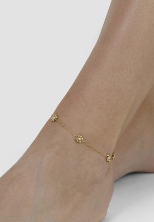 Delicate gold anklet with small flower-shaped charms worn on a bare ankle against white background.