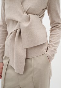 Beige Strickpullover mit drapiertem Vorderteil und seitlichem Knoten-Detail. Leichter Stoff, lange Ärmel und eine taillierte Passform in Kombination mit hellbeigen Hosen.
