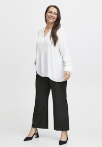 Leende kvinna står med händerna i fickorna, iförd en vit långärmad blus, svarta vida byxor och svarta klackskor.