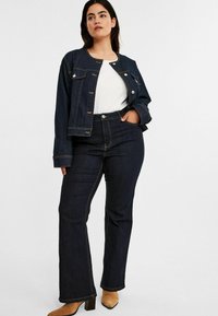 Takataka bulu gelap dengan kerah bulat dan penutupan kancing, dipasangkan dengan celana denim model flare dengan pinggang tinggi yang memiliki jahitan kontras.
