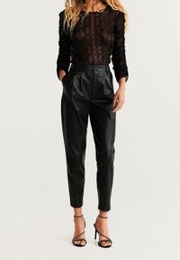 Haut en dentelle noir à manches longues, présentant des motifs complexes ; associé à un pantalon en cuir noir taille haute et des talons noirs à lanières.