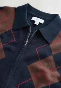 Námornícky zapínací sveter s golierom, ktorý má hnedý geometrický vzor a červené mriežkové akcenty. Vyrobený z pleteného materiálu s hladkou textúrou.