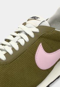 Nike tenisice s gornjim dijelom od maslinasto zelene kordone, bijelim vezicama, ružičastim Swoosh logotipom i kremastim jezikom s logotipom. Crna potplat.