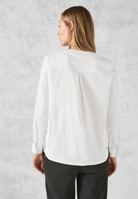 Weißes Langarm-Blusenshirt mit einem Rückenschlitz und knöpfbaren Manschetten, hergestellt aus glattem Stoff. Kombiniert mit dunkelgrauen Hosen, die eine entspannte Passform zeigen.
