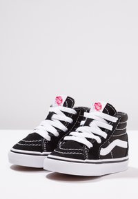 Schwarze Wildleder-Hochtop-Sneaker mit weißen Schnürsenkeln und Sohle. Verfügt über vertikale Nähte und ein rotes Etikett auf der Zunge. Klassisches Design.