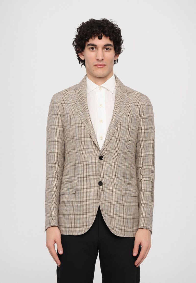 Beige karierter Blazer mit Reverskragen, zwei vorderen Taschen und zwei schwarzen Knöpfen. Darüber ein weißes Hemd getragen und kombiniert mit schwarzen Hosen.