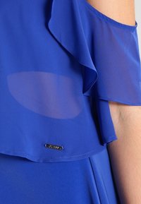 Blusa azul con volantes en capas de tela transparente y opaca, que presenta una pequeña etiqueta metálica con el nombre de la marca en el dobladillo.