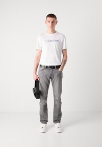 Balta kokvilnas T-kreklā ar zilu "Calvin Klein" logotipu, pelēka džinsu bikses, melna josta un melna soma; modelis, tērpies baltām sporta apavām, stāv taisni.