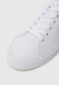 Weiße Sneaker mit einer glatten synthetischen Oberseite, runder Zehenpartie, strukturiertem Gummisohle und flachen weißen Schnürsenkeln. Mit Seitennähten als Detail.