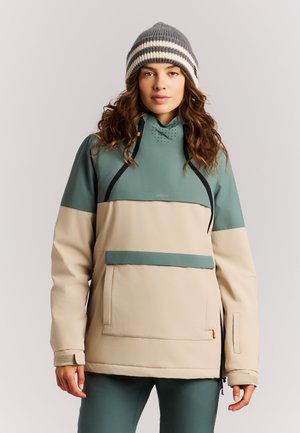 Vrouw met een groene en beige winterjas met zwarte ritsaccenten en een grijs gestreepte gebreide muts, staand voor een effen achtergrond.