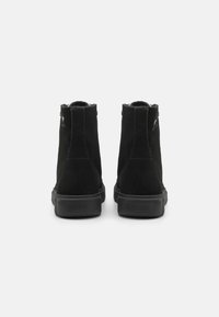 Bottes de cheville en suede noir avec une finition texturée, bout rond et semelles en caoutchouc. Présente des détails de couture à l'arrière et un logo sur le talon.