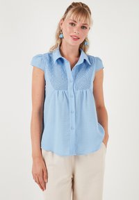 Camicia button-up azzurro chiaro con maniche corte, caratterizzata da ricami testurizzati sul petto, tessuto morbido e orlo curvo.