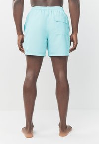 Een paar lichtblauwe zwembroeken met een elastische tailleband en een achterzak, van achteren gezien op blote benen. Glad materiaal met een eenvoudig ontwerp.