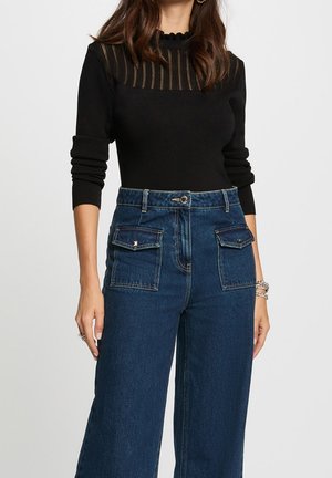 Kvinde iført sort langærmet top med gennemsigtig, stribet yoke og bølget halsudskæring, kombineret med højttaljede blå jeans med forlommer med klap.