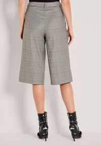 Frau in grau karierten, weit geschnittenen, knöchellangen Hosen und schwarzen, hochhackigen Stiefeletten, die vor einem schlichten, hellen Hintergrund steht.