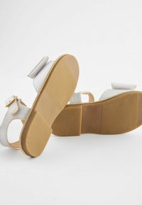 Vita sandaler med en platt, texturerad beige sula. Justerbar ankelrem med ett guldspänne och en dekorativ rosett på den övre remmen.