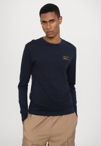 Jūrų laivyno ilgarankovis marškinėliai, pagaminti iš medvilnės mišinio, su apvalia iškirpte, auksiniu logotipo ženkliuku krūtinėje ir šoninėmis siūlėmis.