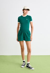 Kvinde iført teal sportsbeklædning, beige hat, hvide sokker og sorte højtop sneakers, stående på grøn gulv mod hvid baggrund.