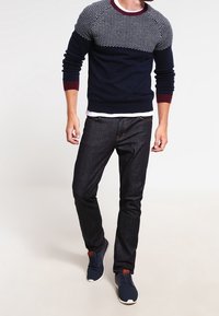 En man bär en marinblå och mönstrad tröja, mörka jeans och sneakers, står mot en ljus bakgrund med en avslappnad hållning.