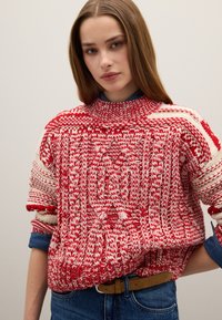Maglione lavorato a maglia rosso e bianco con collo alto, design a trecce prominenti e maniche arrotolate, abbinato a una camicia di jeans blu indossata sotto.