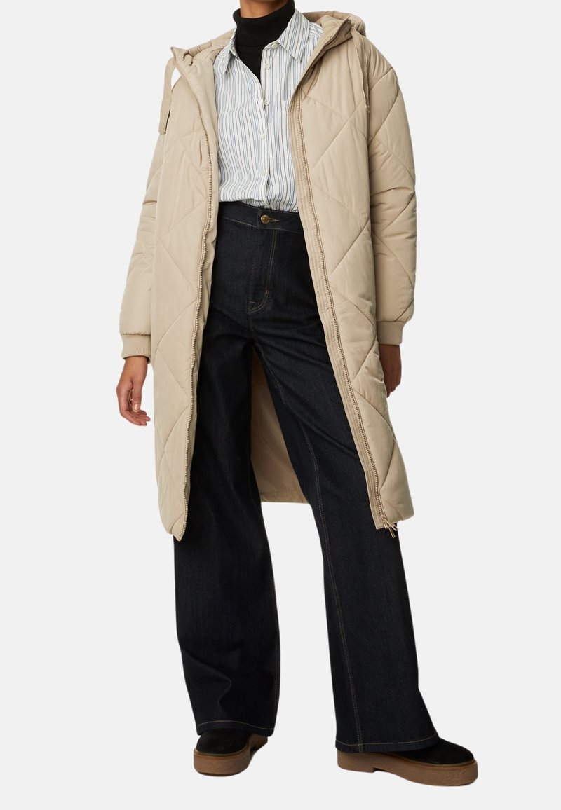 Abrigo largo acolchado beige con capucha, con un detalle de cremallera, combinado con unos jeans de pierna ancha en color azul marino y zapatos negros. Camiseta a rayas debajo.