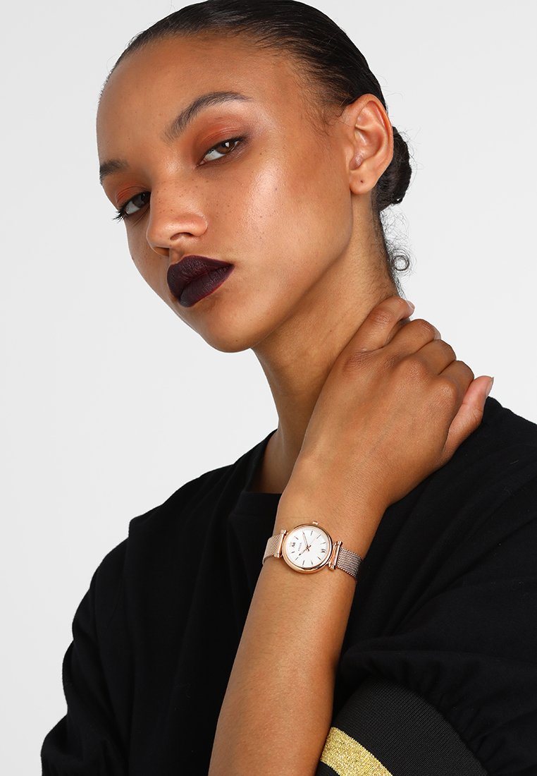Roségoldene Metalluhr mit Mesh-Armband, rundem weißen Zifferblatt und silbernen Akzenten. Das Modell trägt ein schwarzes Oberteil mit Rüschensleeves.