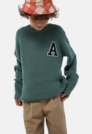 Pull en tricot vert avec une texture côtelée, arborant un patch en forme de lettre "A" marine sur la poitrine. Assorti avec un pantalon beige et un chapeau seau à carreaux.