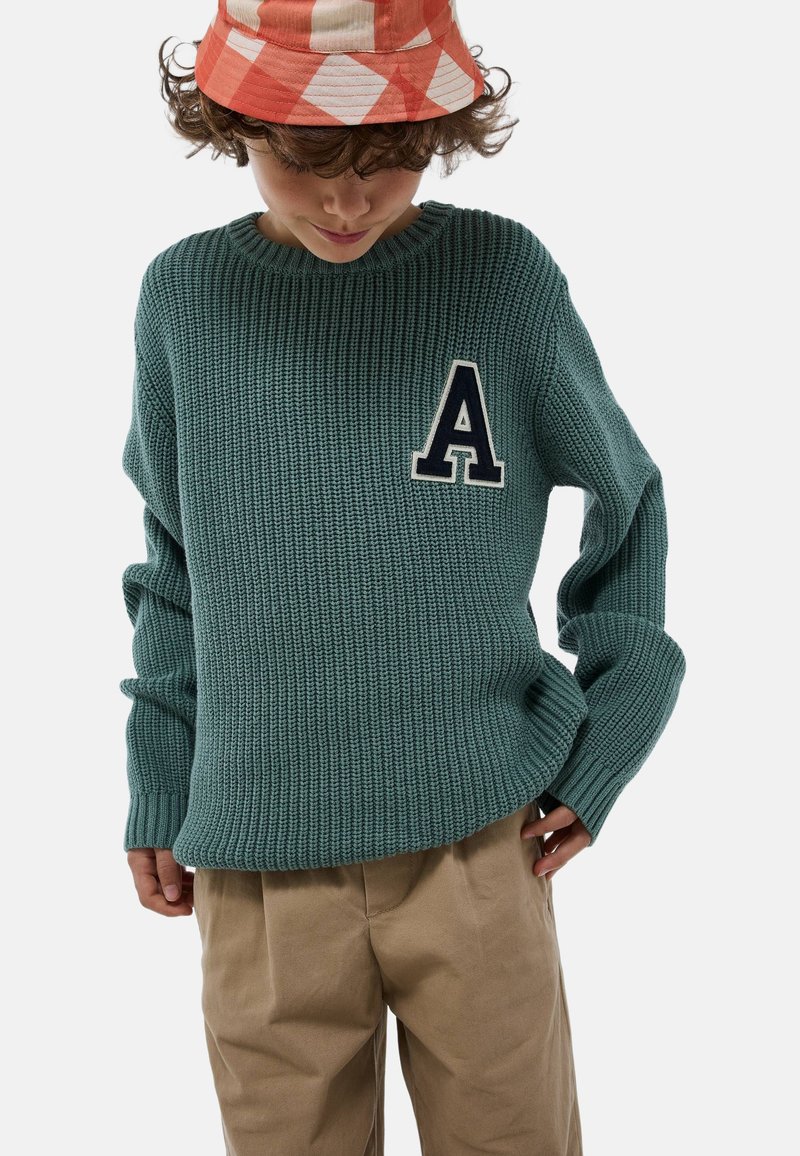 Pull en tricot vert avec une texture côtelée, arborant un patch en forme de lettre "A" marine sur la poitrine. Assorti avec un pantalon beige et un chapeau seau à carreaux.