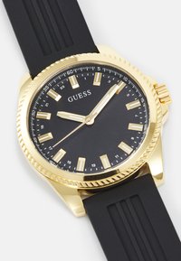Montre à cadran noir avec un boîtier en or, des marqueurs dorés et une lunette rotative. Elle est dotée d'un bracelet en caoutchouc noir avec des accents en rainure pour ajouter de la texture.