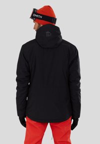 Černá voděodolná bunda s kapucí, s zipovým uzávěrem a elastickými manžetami, doplněná o jasně oranžové kalhoty a pletenou čepici.