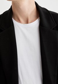 Svart blazer med ett herringbone-mönster, bärs över en vit t-shirt med rund hals. Fokusera på kragen och axlarna på blazern.