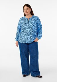 Blusa a maniche lunghe azzurro chiaro con motivo astratto nero, scollatura rotonda e chiusura con bottoni. Abbinata a jeans in denim blu a gamba larga.