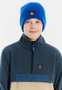 Blauer Strickbeanie und ein Fleece-Oberteil mit einem Reißverschlusskragen, dunkelblauem oberen Bereich, beigem Unterteil und blauem Logostick. 